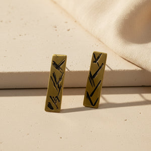 A pair of gold-colored elongated brass earrings with a signature 'X' texture and a deep patina finish.