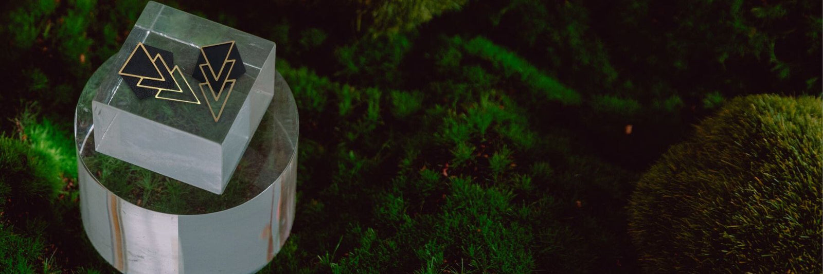 Earrings in upcycled leather and brass are sitting atop an acrylic block over grass.