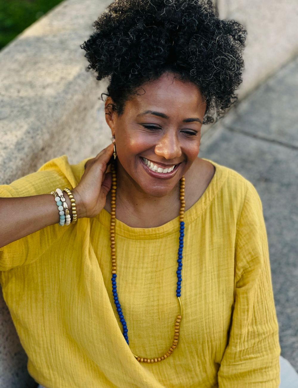 A woman is wearing a yellow top with a beaded wrap bracelet and long beaded necklace.